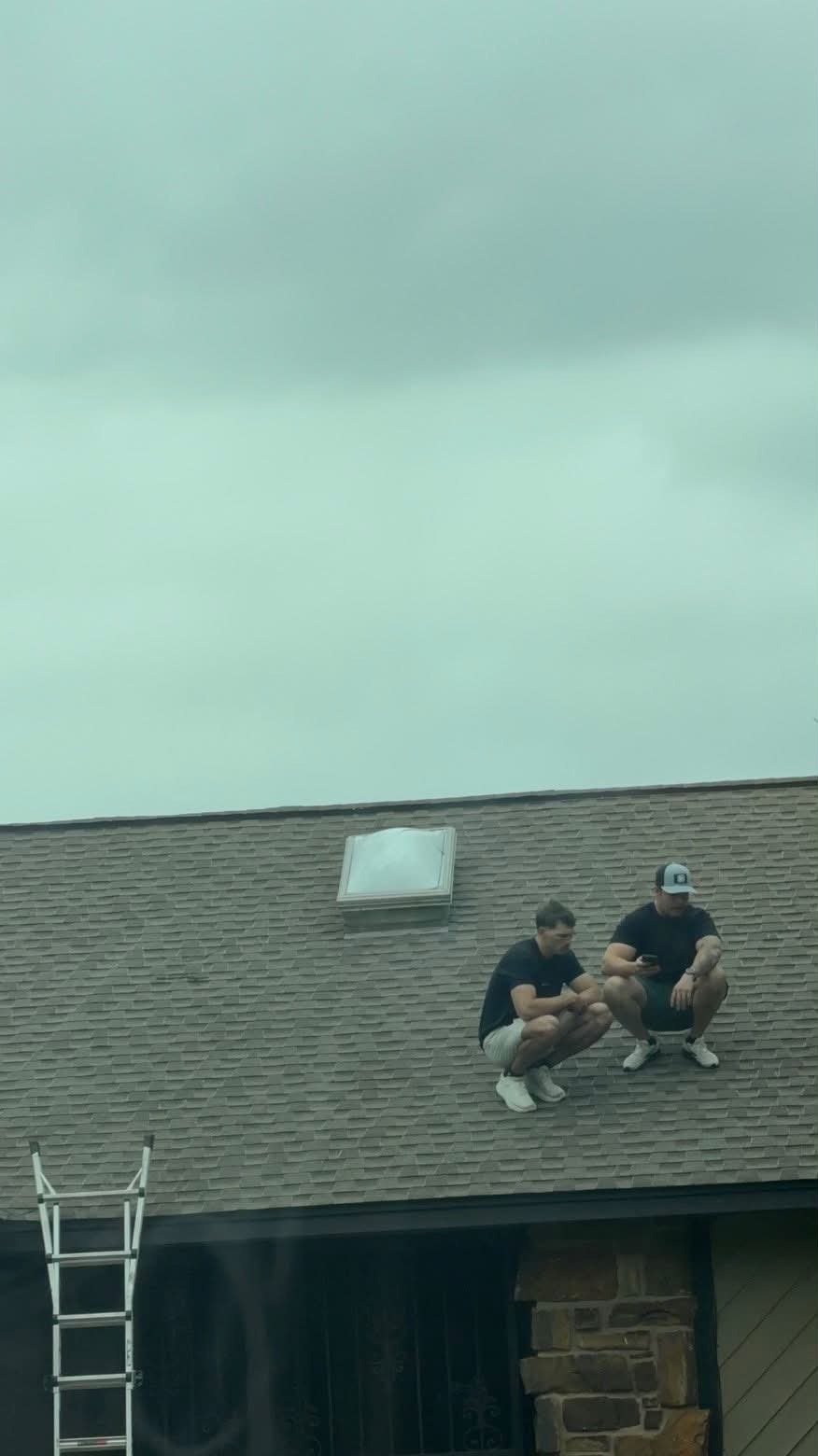 Two Aces Roofing and Exterior Solutions contractors crouch on a gray asphalt shingle roof inspecting a broken skylight dome in Verdigris, Oklahoma, with an extension ladder leaning against the structure below.
