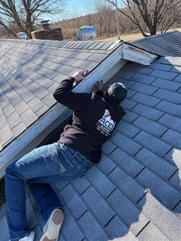 A roofing professional from ACES Roofing and Exteriors Solutions inspects a residential roof on Bluff Rd, Claremore, OK (GPS: 36° 23' 51.75" N, 95° 34' 42.95" W; Altitude: 220m). The inspector is prone on grey asphalt shingles near several silver turbine vents and a brick chimney, examining a gabled section where the white wooden trim shows weathered, peeling paint.
