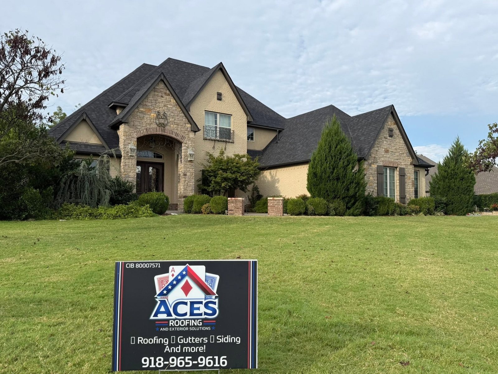 Large stone and stucco two-story home in Stone Canyon, OK with new Class 3 Owens Corning Duration roof in Onyx Black, Aces Roofing yard sign in foreground