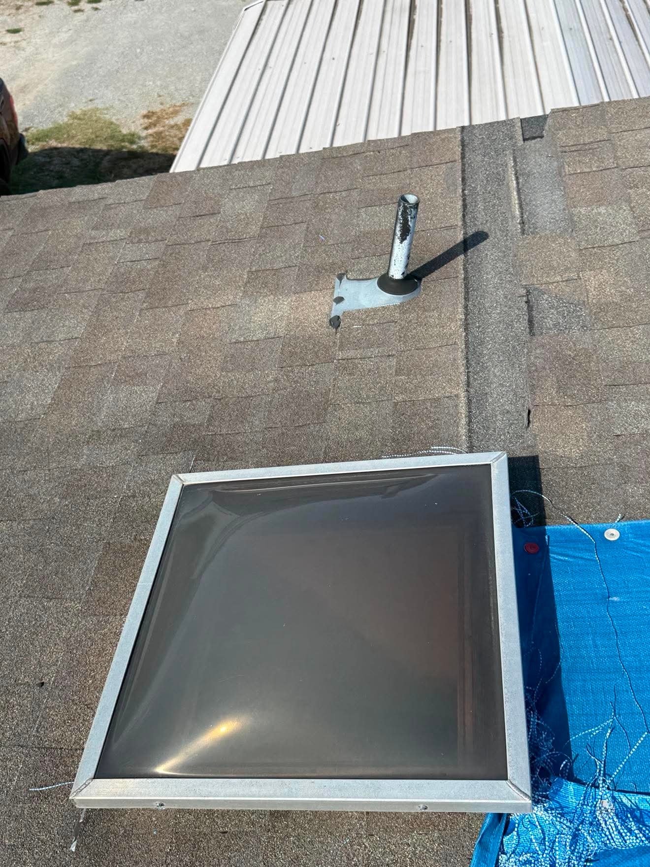 An Aces Roofing and Exterior Solutions crew stages a replacement flat-glass skylight unit with an aluminum frame on a brown asphalt shingle roof during a skylight repair in Inola, Oklahoma, with a pipe flashing vent and blue tarp visible nearby